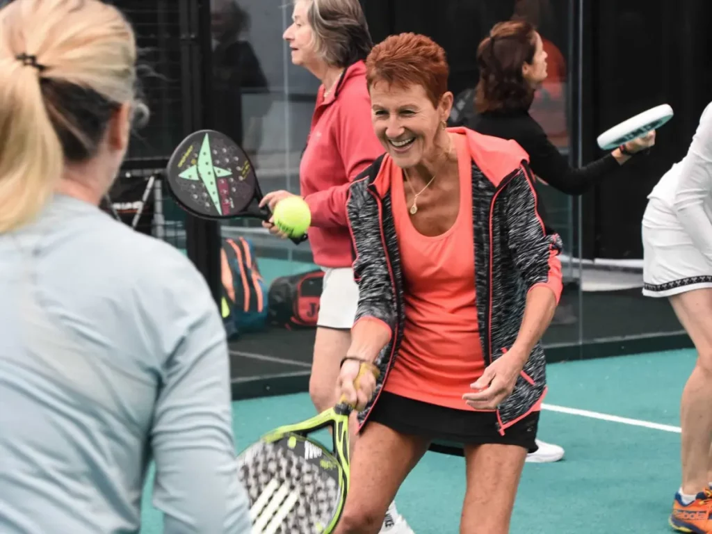 Female padel player with partner doing warm up on court, with lots of players in similar space practicing