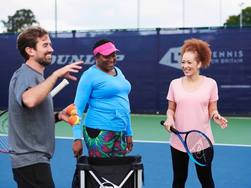 Coach with 2 female players. Coach is stood in front of ball basket, with the 2 women smiling and listening