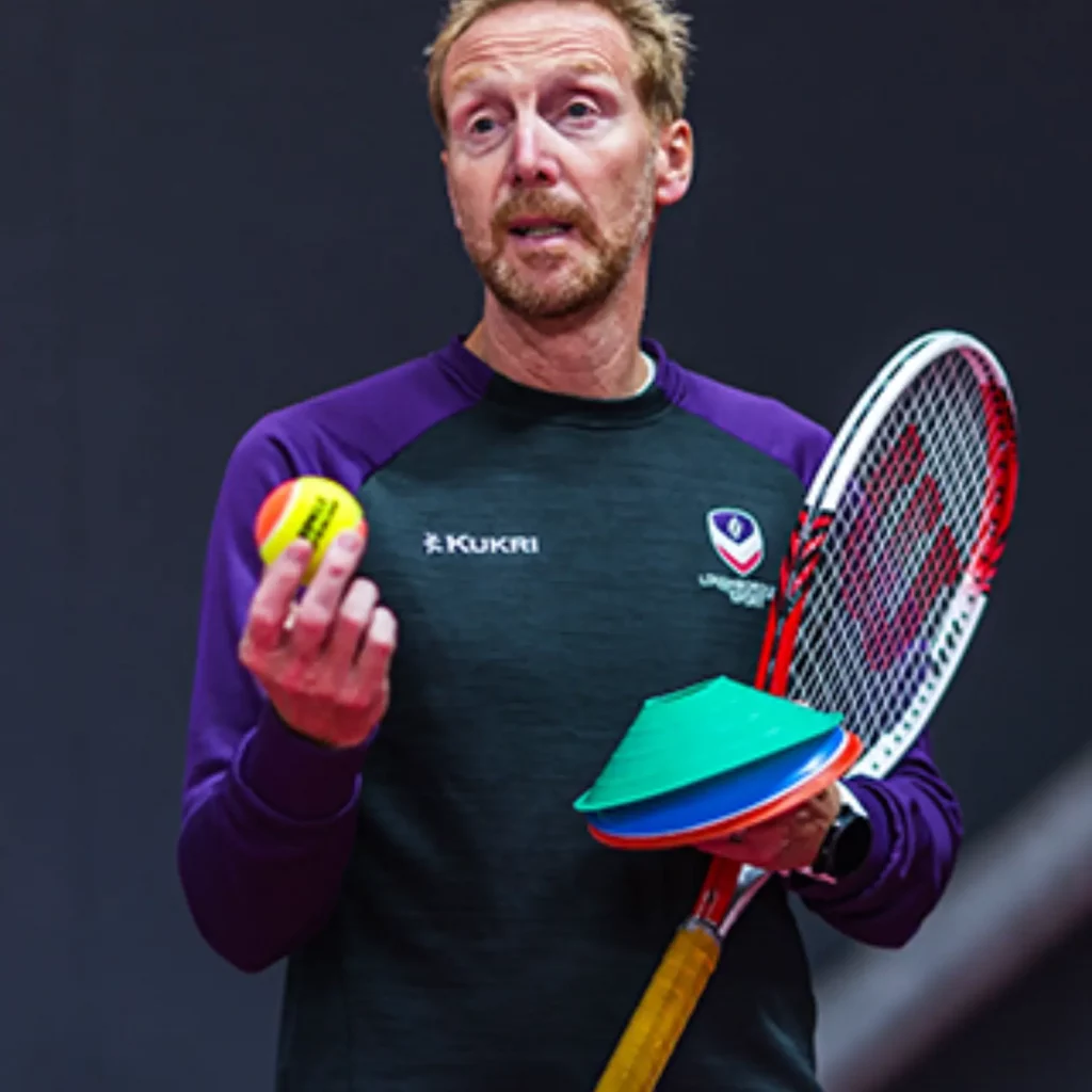 Dave holding tennis ball in right hand and cones / racket in other and sharing info to coaches