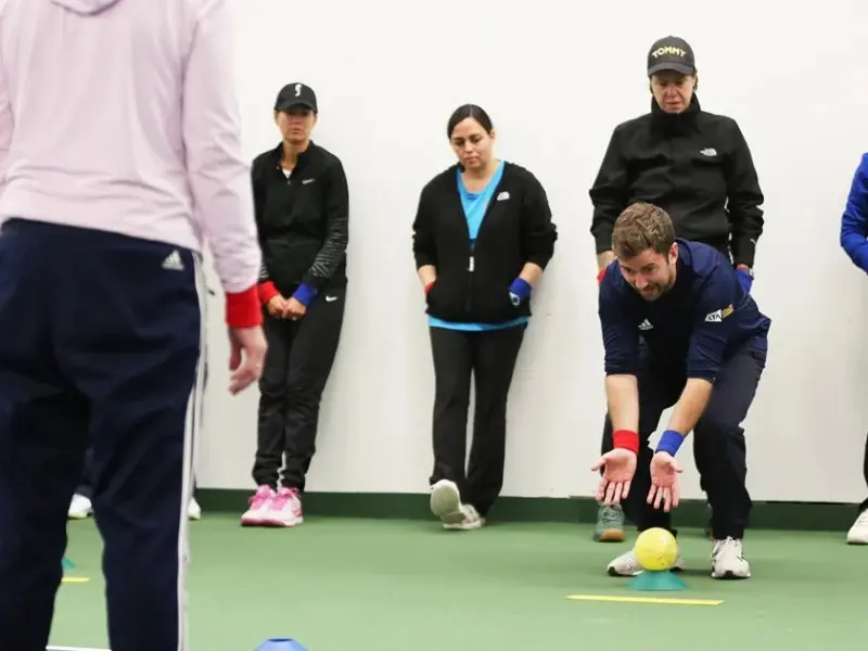 Coach rolling bright yellow ball to a partner, with people stood watching the demonstration