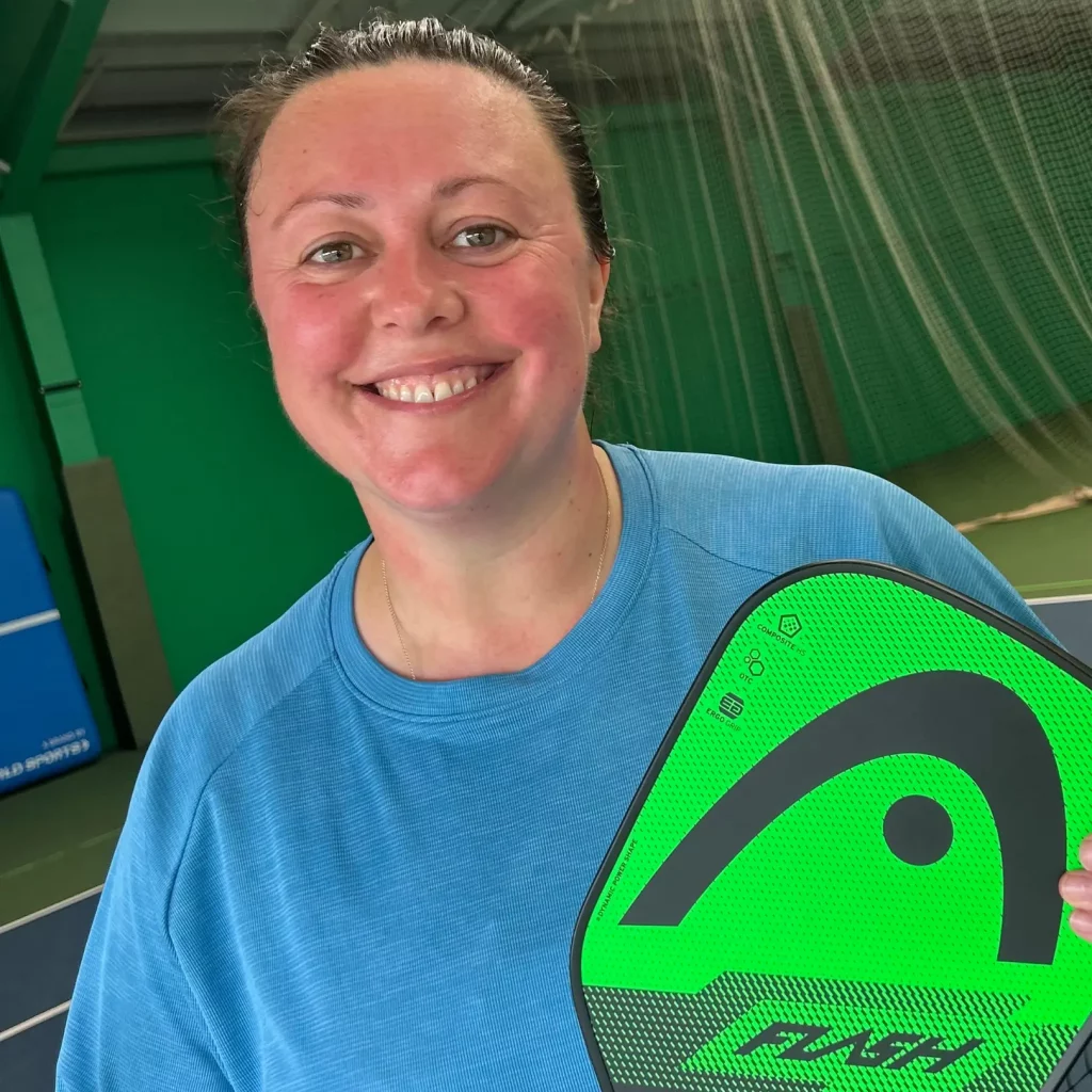 Leah, holding pickleball racket, with hair tied back and smiling at the camera