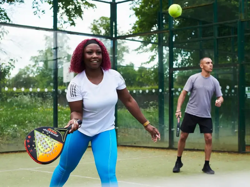 Woman with blue leggings and white t shirt hitting a padel ball with multicoloured racket, with partner in background