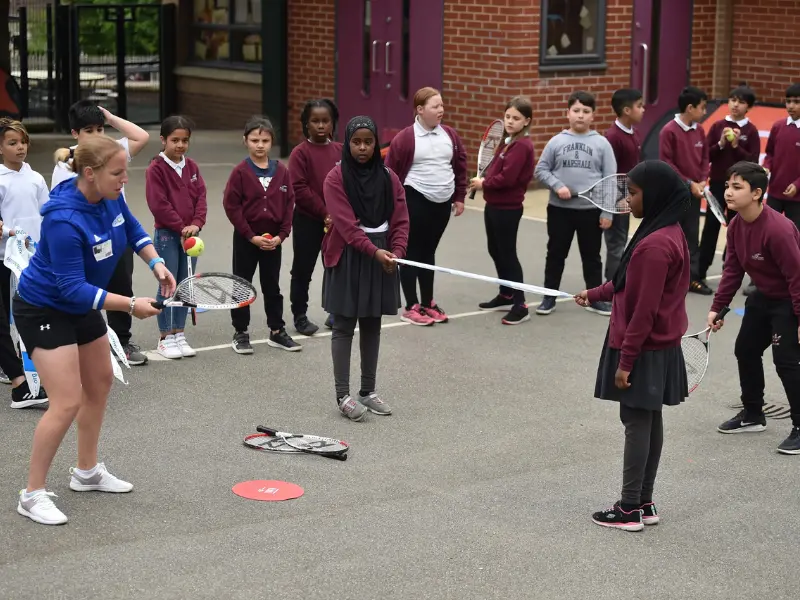 Ang showing school children how to hit ball over barrier tape, being held by two pupils.