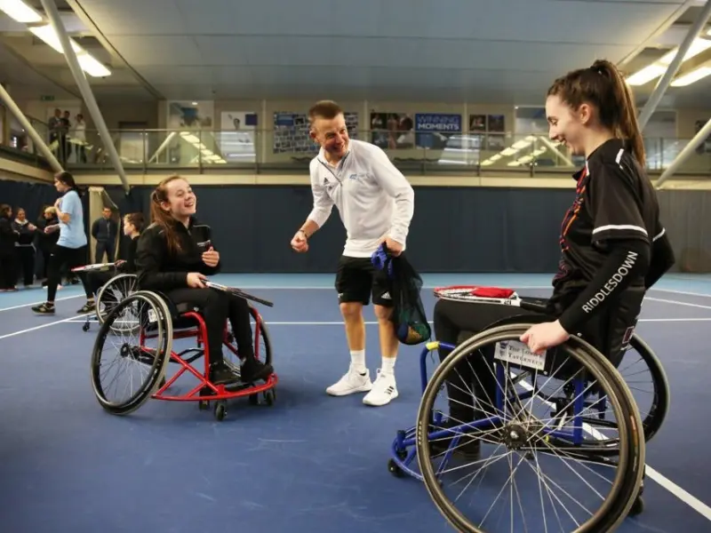 Male coach in white jacket in middle of wheelchair players, ready to play tennis.