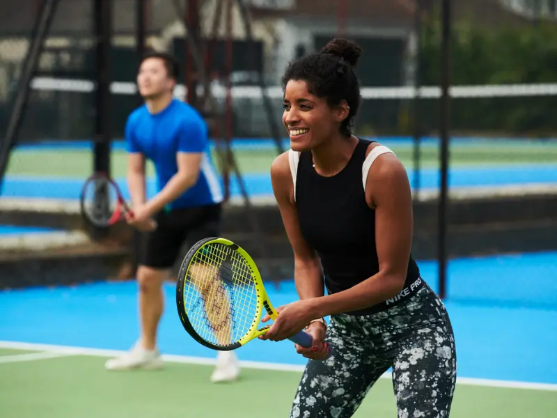 Female player stood at net with racket out in front, with male player behind at the baseline, also in ready position