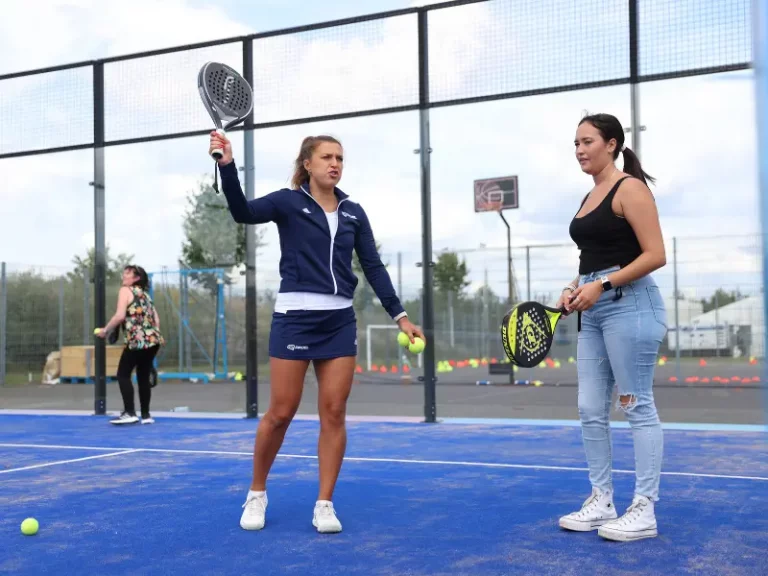 Female padel coach demonstrating an overhead, to a female player listening and ready to play